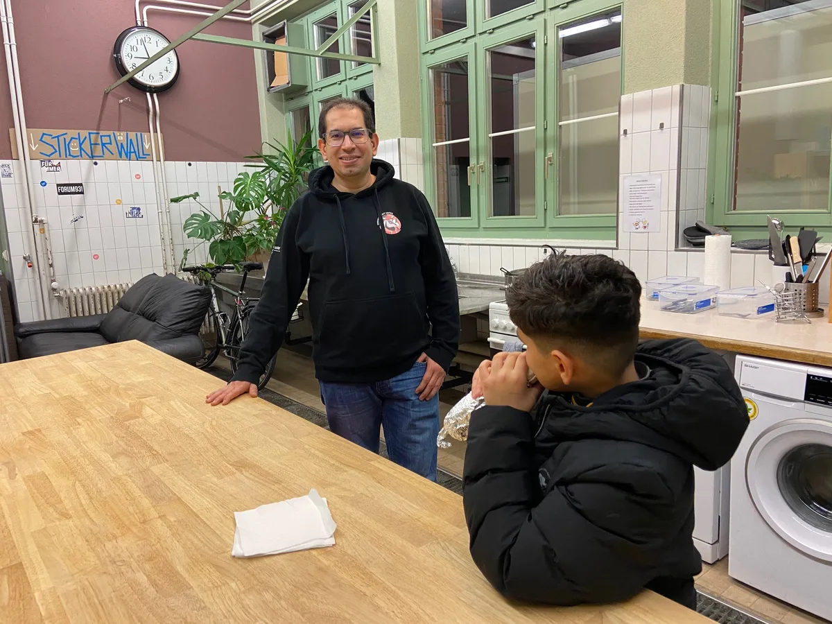 Der Junge hat Hilfe bekommen von Mohammad Majdalawi, der durch seine Sprache einfachen Zugang findet. Ein erwachsener Mann stützt sich auf einen Tisch, davor sitzt ein kleiner Junge und isst einen Kebab.